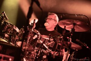 Drummer on stage playing drums under red lighting, blowing a bubble while performing live