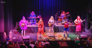 Full-stage view of a seven-member band performing live in vibrant costumes under blue-purple lights, with an audience at tables in the foreground.