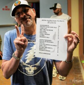 Man in a skull-print shirt and baseball cap holds up a printed list of songs, smiling at the camera, making a peace sign.