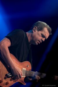 Guitarist in a black shirt plays a sunburst electric guitar on stage with blue stage lighting in the background.