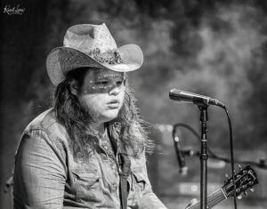 Musician wearing a cowboy hat and denim shirt performs on stage with an electric guitar and microphone in front of him.