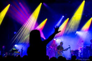 Crowd raising hands at a concert with a band onstage and bright multicolored spotlights (blue, purple, yellow).