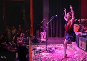 Female singer on stage raises both arms, microphone in hand, under pink stage lights with an excited audience nearby.
