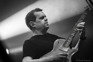 Male guitarist on stage playing an electric guitar, close-up side profile with guitar neck in frame (black and white).