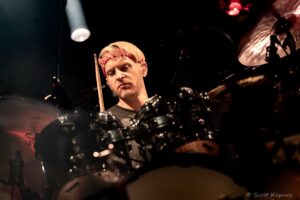 Drummer with blond hair and a red headband playing drums on a dimly lit stage.