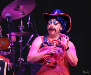 Performer in a blue wig and red sequined hat with stage makeup, holding a prop accessory on a dimly lit stage with drums in the background