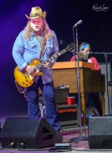Guitarist in a denim shirt and cowboy hat playing a yellow Les Paul on stage with purple lighting; keyboardist in background.