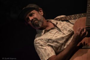 Male guitarist wearing a hat and floral shirt plays an acoustic guitar on a dark stage.