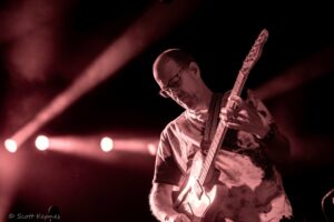 Guitarist wearing glasses playing an electric guitar on stage bathed in red lights.