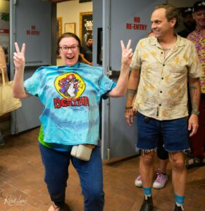 Smiling woman in a blue tie-dye shirt and glasses raises a peace sign beside a man in a beige patterned shirt, both posing for a photo at a doorway.
