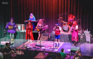 A lively band performs on a stage with colorful costumes and instruments in front of an audience.