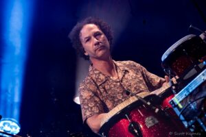 Male musician in a floral shirt playing a glittery red drum set on stage under blue lights.