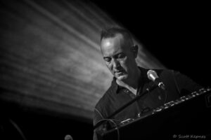 Black-and-white photo of a man performing on stage, singing into a microphone while playing a keyboard on a dimly lit backdrop.