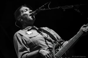 Black-and-white photo of a male bassist singing into a microphone on stage, performing live