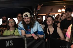Crowd of concertgoers leaning on a barrier, some smiling, a man in a cap making horn gesture toward the stage in a dim venue: close-knit group at a show.