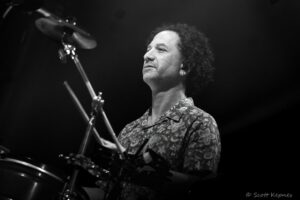 Black-and-white photo of a smiling drummer playing a drum kit on stage, cymbals visible in the foreground.