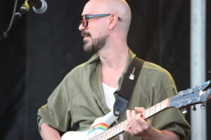 Male guitarist with shaved head and beard wearing sunglasses, playing a white electric guitar on stage.