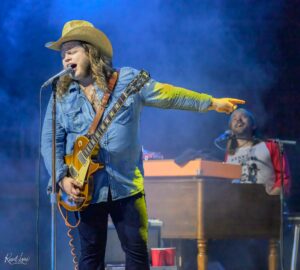 Male guitarist and singer in a denim shirt and cowboy hat performs on stage, microphone in front, blue stage lighting.
