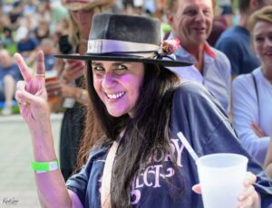 Smiling festival attendee wearing a black hat with a ribbon and flower, waving a peace sign at the camera
