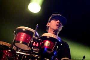 Close-up of a drummer wearing a cap and glasses, playing a glittery red drum set under a bright yellow stage light during a concert.