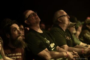 Audience members leaning on a barrier at night, looking up toward the stage as they watch a performance.