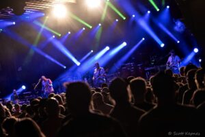 Crowd watches a live band on a dimly lit stage with blue and green spotlights sweeping overhead.