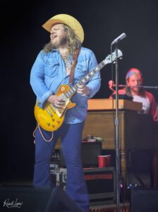 Guitarist in a denim shirt and straw hat playing a yellow Les Paul on stage, eyes closed in performance.