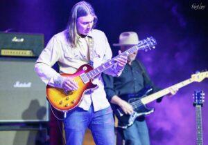 Guitarist playing a sunburst Les Paul on stage with purple lighting and amplifiers in the background.