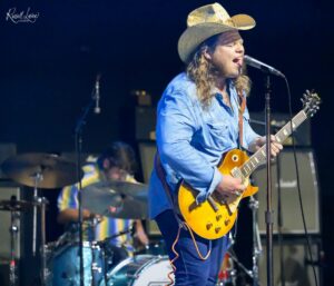 Guitarist in a blue shirt and cowboy hat sings into a microphone on stage.