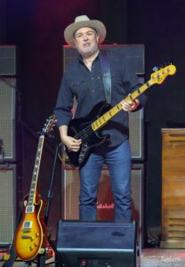 Male guitarist wearing a wide-brimmed hat and dark shirt playing a black electric bass on a stage with amplifiers in the background.