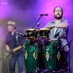 Musician with glasses and beard playing green conga drums on a stage with purple lighting and a microphone nearby.