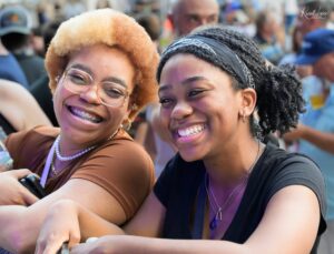Two women posing for a selfie at a crowded outdoor event, smiling at the camera; one with blond curly hair and glasses, the other with a black bandana and braided hair.