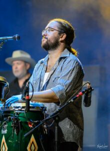 Musician with glasses and beard, wearing a checkered shirt, playing percussion on stage with blue lighting in the background.