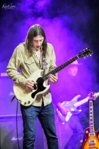 Guitarist in a beige shirt playing a white Gibson SG on stage with purple lighting and a smoky backdrop; another guitar visible in the background.