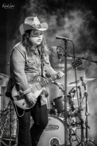Guitarist wearing a cowboy hat and denim shirt performing on a smoky stage with a microphone nearby.