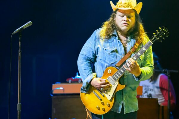 Male guitarist wearing a yellow cowboy hat and denim shirt, playing a sunburst Les Paul on stage with a mic stand nearby.