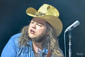 Male musician with long hair wearing a wide straw hat on stage, guitar strap visible, microphone nearby.