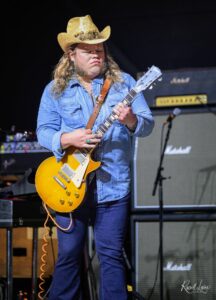 Male guitarist wearing a straw hat and denim shirt, playing a yellow Les Paul on stage with Marshall amps in the background.