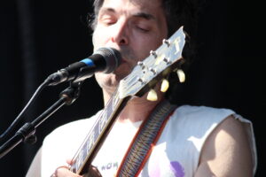 Male musician singing into a microphone while playing an acoustic guitar on stage, close-up of face and guitar headstock with tuning pegs visible