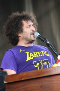 Male singer with curly hair wearing a purple Lakers shirt, singing into a microphone on stage.