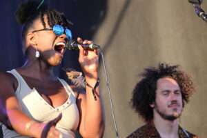 Female singer wearing blue mirrored sunglasses, singing into a handheld microphone on stage with a guitarist nearby.