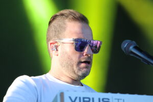 Male singer wearing clear-framed sunglasses sings into a microphone on a stage with a bright lime background.