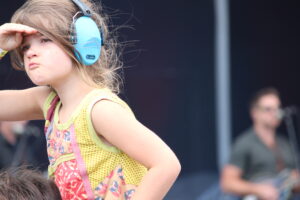 Girl wearing bright blue over-ear headphones, shielding her eyes with one hand during an outdoor event.