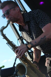 Male saxophonist wearing sunglasses and a patterned shirt plays on an outdoor stage.