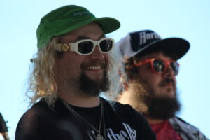 Two men outdoors wearing hats and sunglasses; foreground man with white frames and a green cap smiles, blue sky behind them.