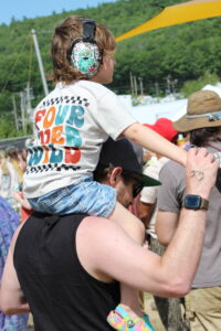 Child sitting on an adult's shoulders at an outdoor event, wearing colorful sneakers and patterned headphones with a cartoon character on them.