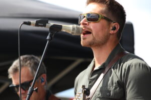 Male singer wearing green shirt and yellow-green sunglasses singing into a microphone on stage outdoors.