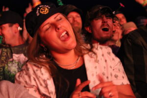 Young woman with a nose ring singing along at a crowded night concert, eyes closed.