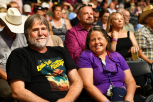Smiling older couple seated at an outdoor event, crowd in the background, man in black shirt and a graphic tee, woman in purple with glasses hanging from her neck.