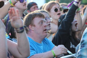 Crowd of excited festival-goers, man in a blue shirt in foreground looking up, cheering with wristbands visible on wrists.